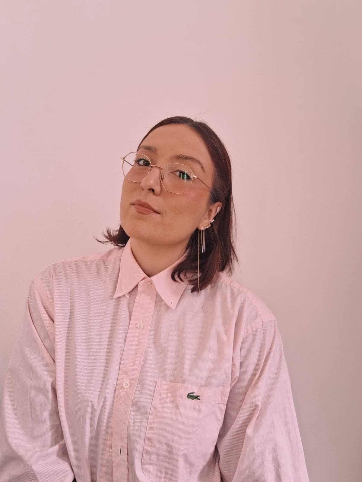 Close-up of a woman wearing a loose-fitting pink shirt with a Lacoste logo on the pocket. She has short hair and round, gold-framed glasses. She wears long, dangling earrings and wears makeup. The woman looks straight into the camera.