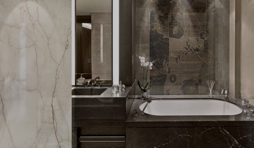 A close-up view of a sleek, modern bathroom. On the left, a white marble panel with dark grain. In the center, a double sink in dark marble. On the right, a white bathtub set into a dark marble cabinet. The back wall is decorated with panels that alternate wood and grainy textures, with a large golden floral motif.