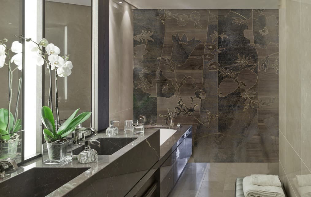 A luxury bathroom with a double sink in dark marble and a white sunken tub. The back wall is covered with large vertical panels that alternate a dark wood-like texture and grainy panels, with a large golden floral motif superimposed. Potted white orchids are positioned on the edge of the sink. The rest of the walls and the floor are made of light marble.
