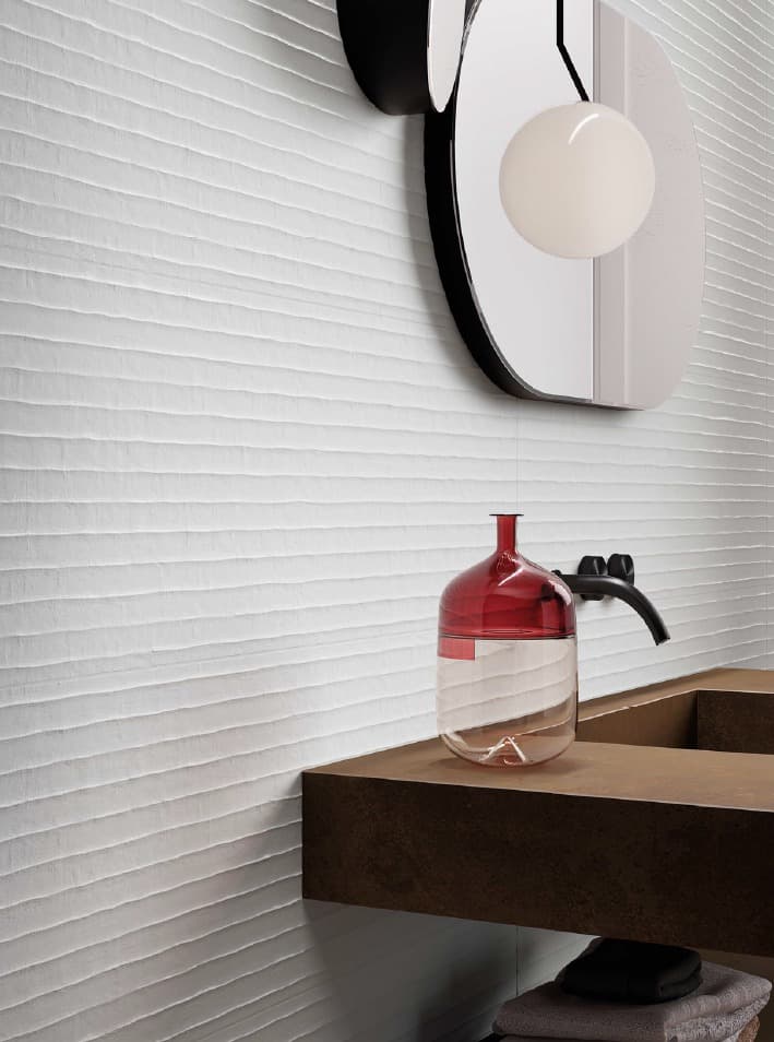 A close-up of a modern sink. The back wall is covered with white relief tiles that create a horizontal wavy pattern. The sink is a single slab of brown stone extending from the wall. A matte black faucet is installed directly on the sink. A red and clear glass vase sits on the edge of the sink. On the wall, an irregularly shaped designer mirror reflects a sphere-shaped lighting.