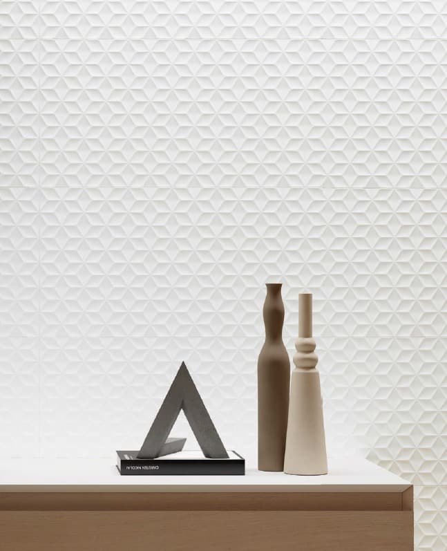 A view of a modern light wood table against a white wall in the background. The wall is covered with three-dimensional white tiles featuring a geometric pattern of raised triangles and cubes. On the table, two elongated ceramic vases, one brown and one white, are placed next to an abstract concrete sculpture in the shape of an "A".
