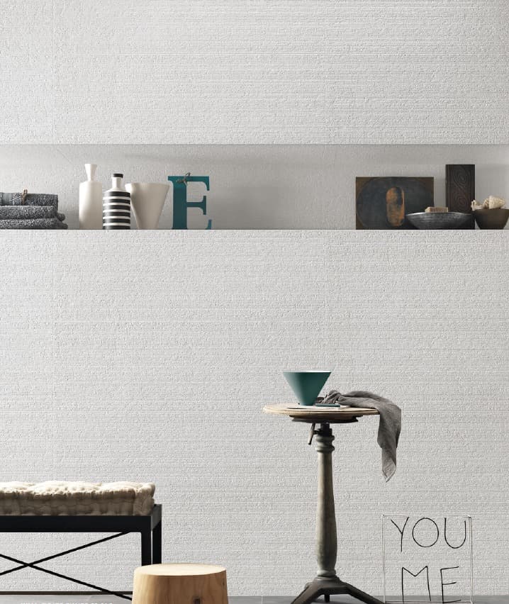 An image of a white wall with a textured, textured effect reminiscent of a tightly woven fabric. The upper section features a horizontal recessed niche, used as a shelf for decorative objects such as vases, books, bowls, and the blue metal "E" sculpture. The lower section of the image shows a shaggy fabric bench, a small cylindrical wooden stool, and a black coffee table with a green bowl. An art installation forming the words "YOU ME" is in the foreground.