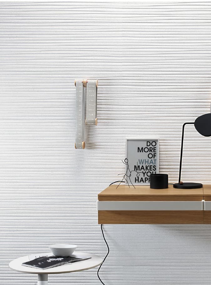 A white wall with a textured, textured effect reminiscent of a wavy fabric. A wooden and fabric calendar hangs in the center of the wall. In the foreground, a modern light wood and white desk is partially visible, with a black table lamp and a small painting reading "DO MORE OF WHAT MAKES YOU HAPPY."
