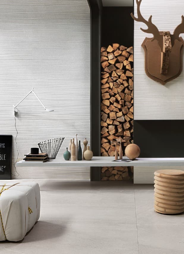 A view of a modern living room with a large wall of white tiles with a horizontal relief pattern. In the center of the room, a recess in the wall is filled with stacks of firewood, serving as decoration. A long white shelf protrudes from the wall, supporting vases and other decorative objects. A white ottoman is positioned on the left side, while a cylindrical wooden stool is on the right. A cardboard deer head hangs on the wall to the right.