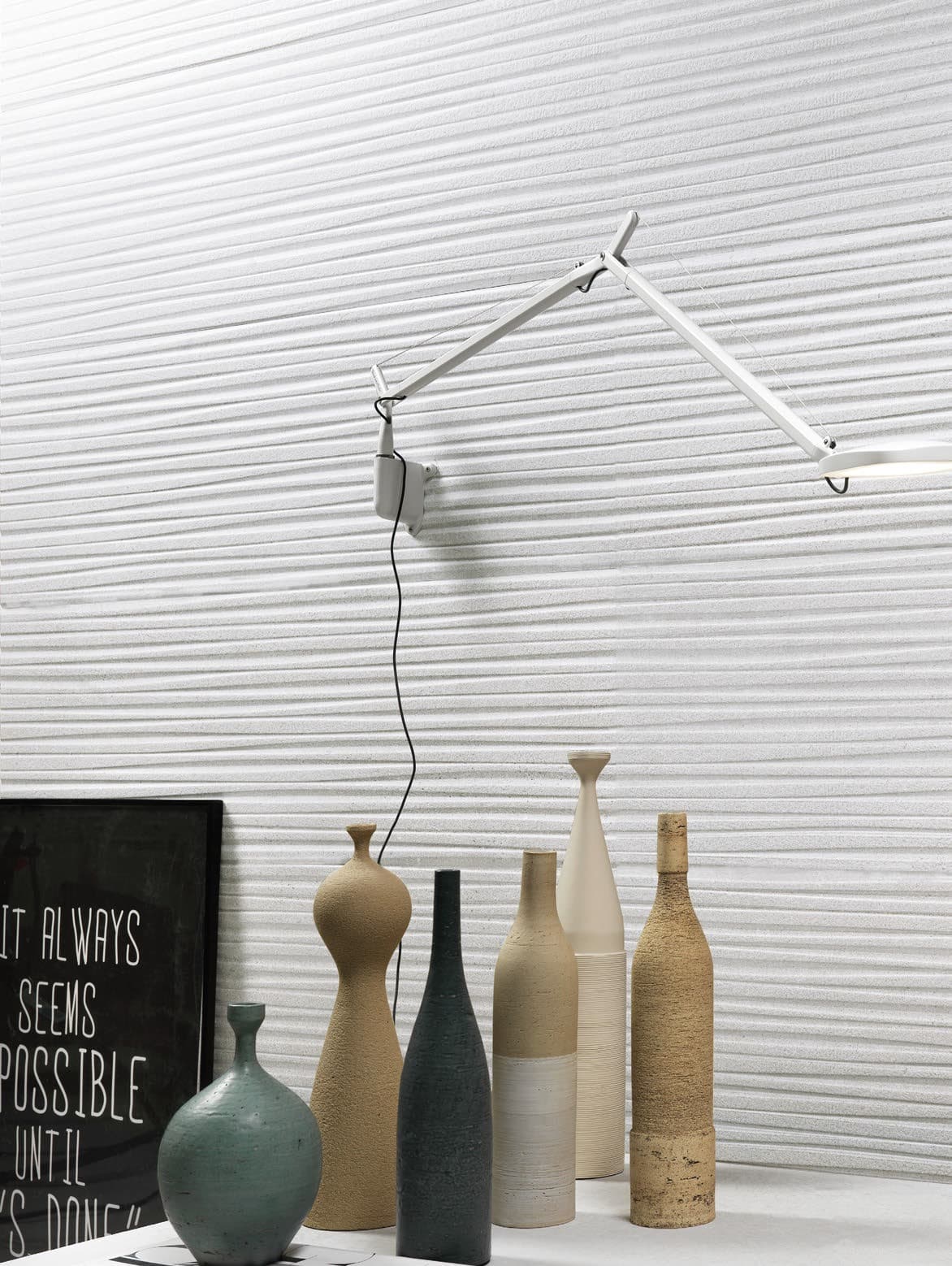 A close-up view of an interior. The back wall is covered with white relief tiles with a,wavy pattern that creates a horizontal striped effect. A white wall lamp with a swivel arm is mounted on the wall. Below, on a white shelf, several ceramic vases of various shapes and heights, in shades of beige, brown, and blue-green, are arranged in a row. On the left side, a black frame with text in white lettering reads: "IT ALWAYS SEEMS IMPOSSIBLE UNTIL IT'S DONE."